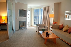 Habitación de hotel con sofá, mesa de centro, televisor, chimenea y un ventanal con cortinas.