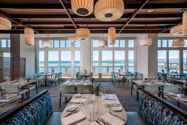 Un restaurant avec des tables dressées pour le service, de grandes fenêtres offrant une vue sur la mer et des lampes suspendues décoratives.