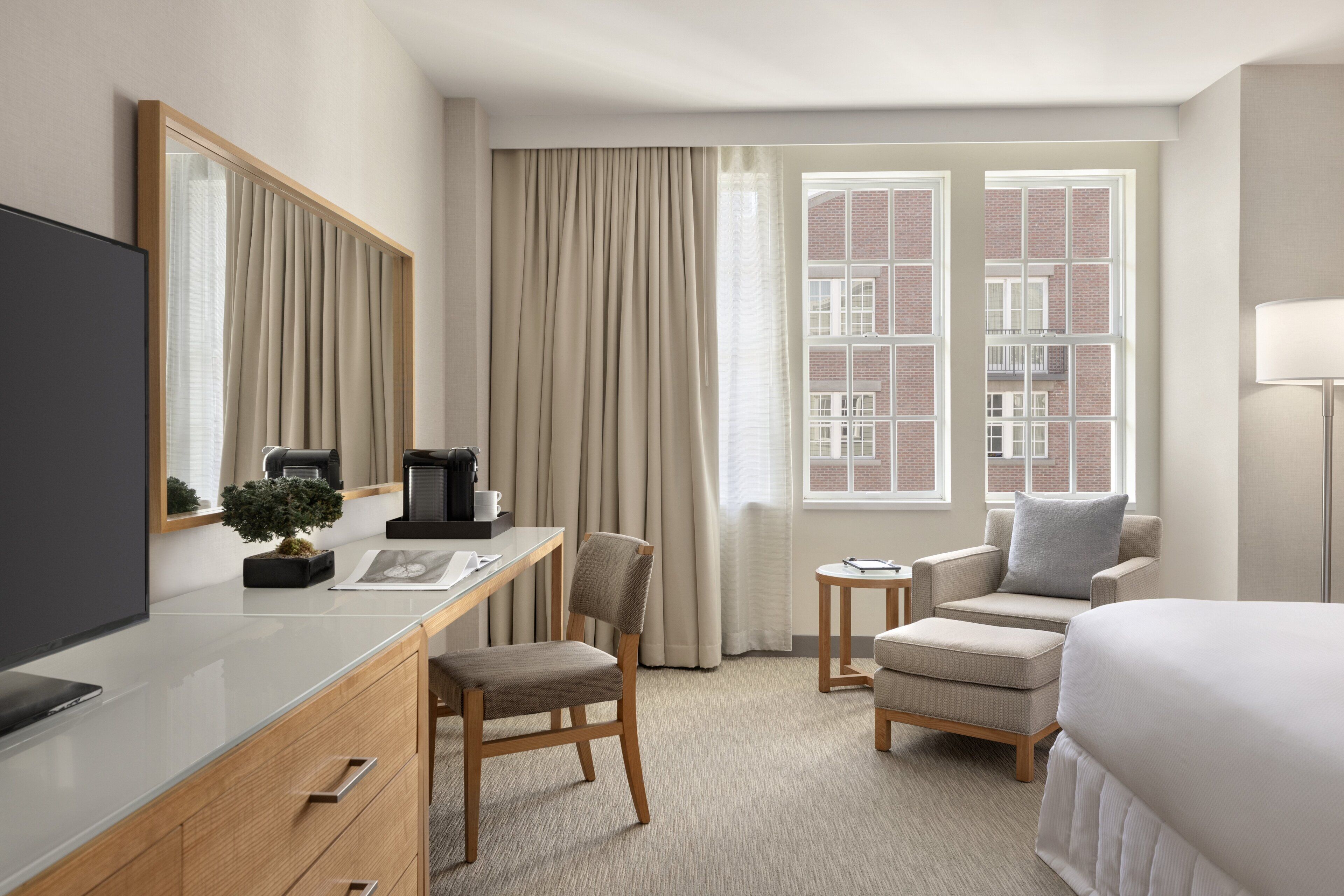 A hotel room with a bed, a desk with a computer, a chair, a small table with a lamp, and a window with curtains.