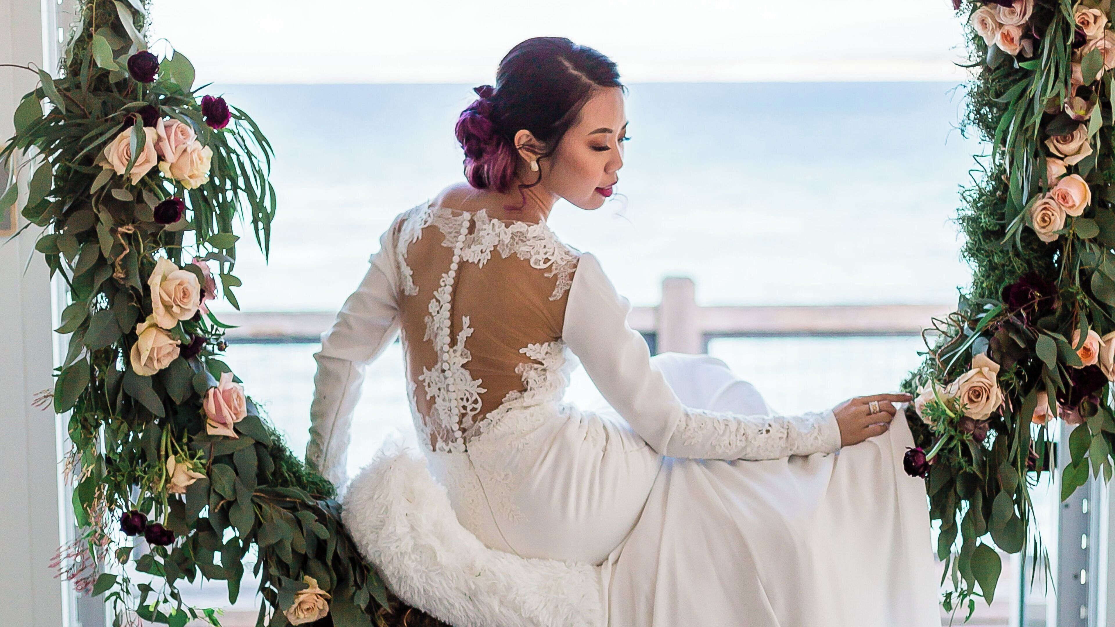 A person in a white dress with lace details, seated by a window with a view of the sea.