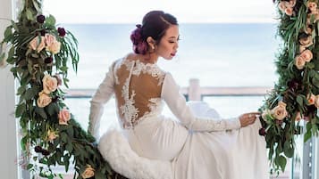 A person in a white dress with lace details, seated by a window with a view of the sea.