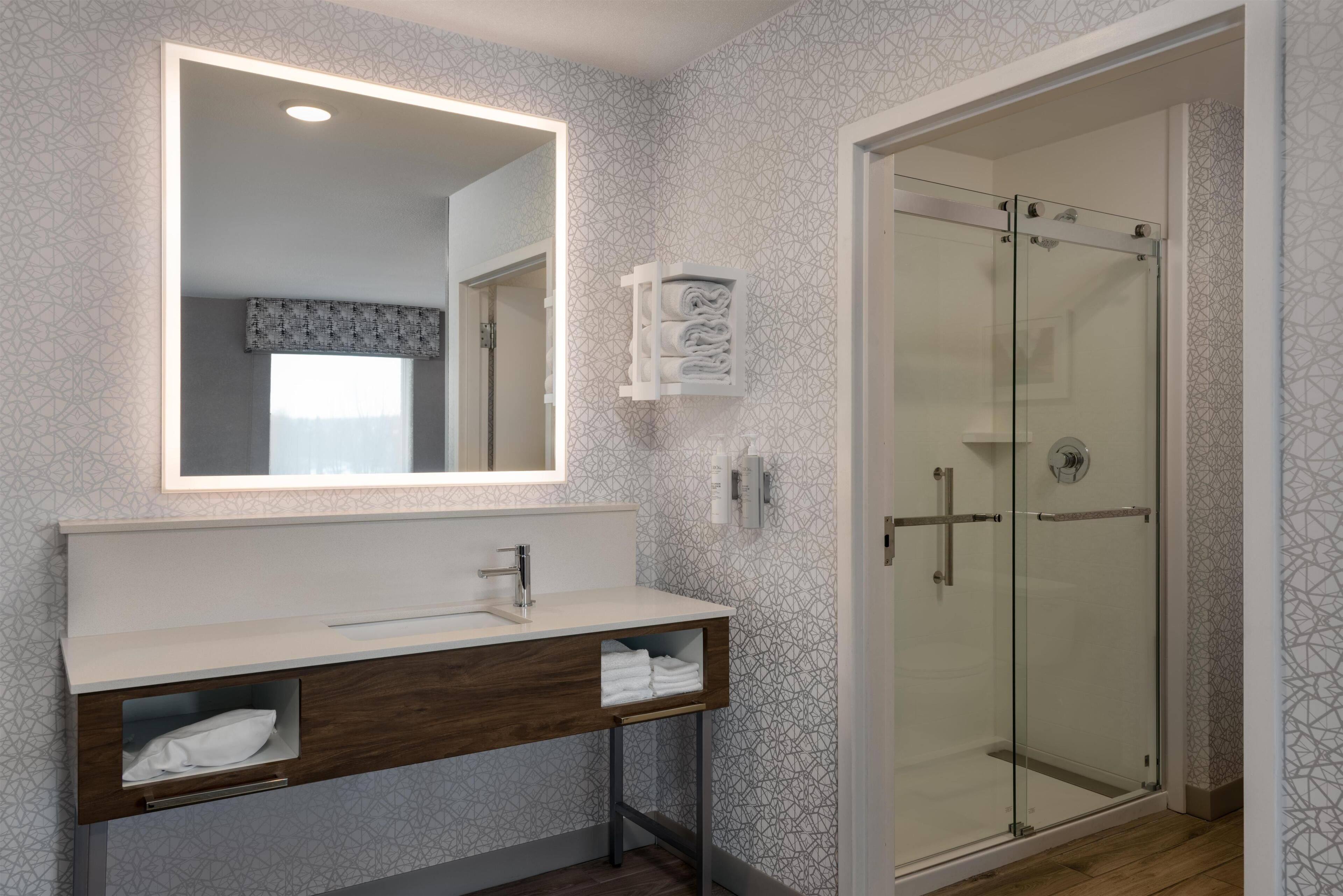 A modern bathroom with a large mirror, a vanity with a sink, a glass shower enclosure, and patterned wallpaper.