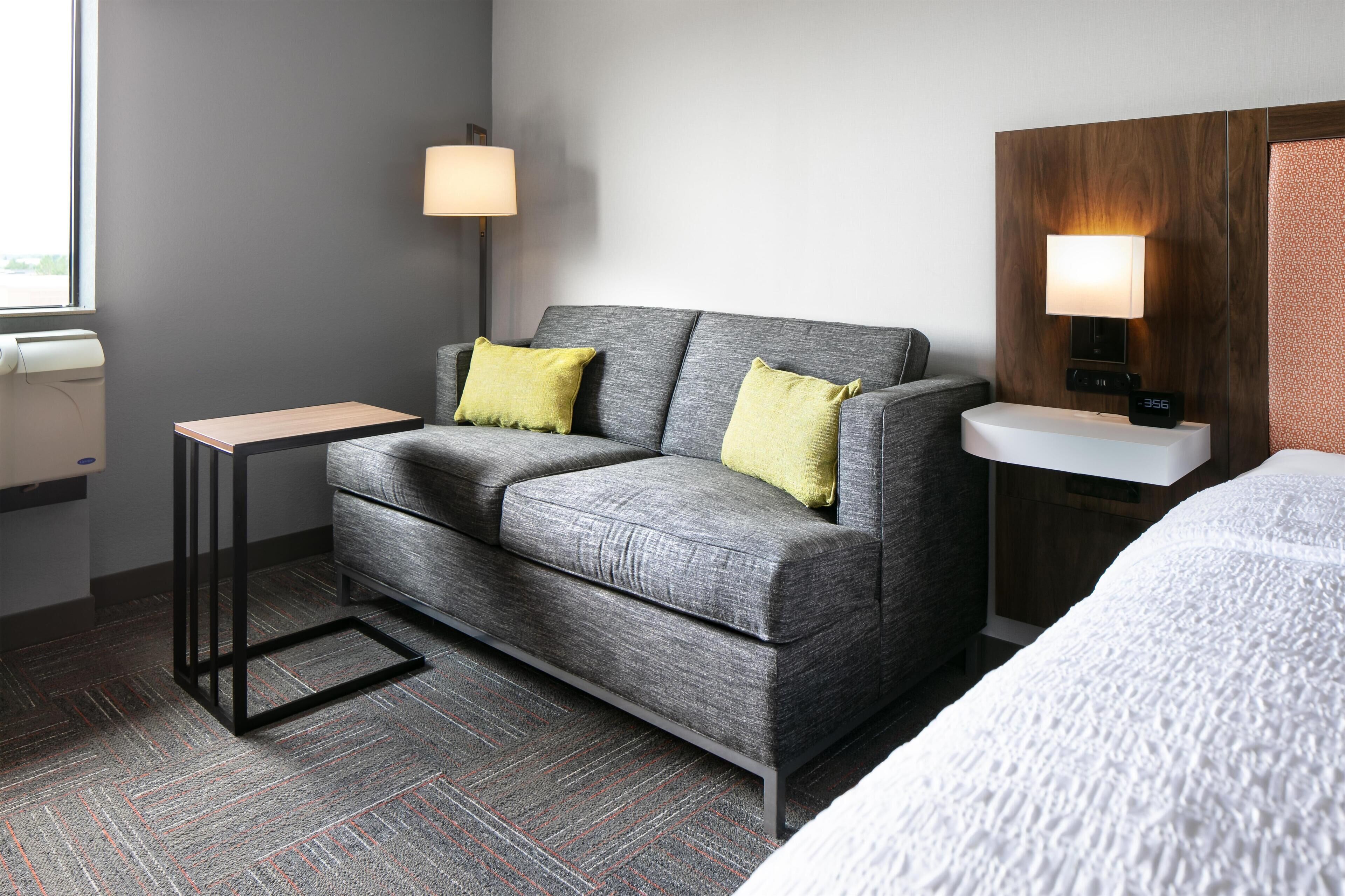 A hotel room with a grey sofa, two yellow pillows, a small table, a bedside lamp, a clock, and a bed with a white cover.