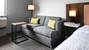 A hotel room with a grey sofa, two yellow pillows, a small table, a bedside lamp, a clock, and a bed with a white cover.
