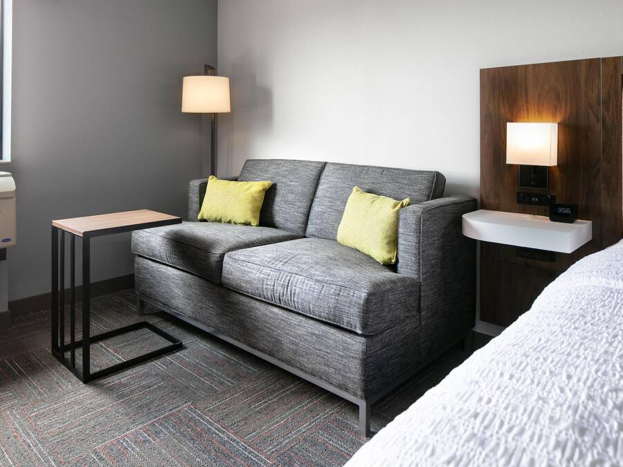 A hotel room with a grey sofa, two yellow pillows, a small table, a bedside lamp, a clock, and a bed with a white cover.