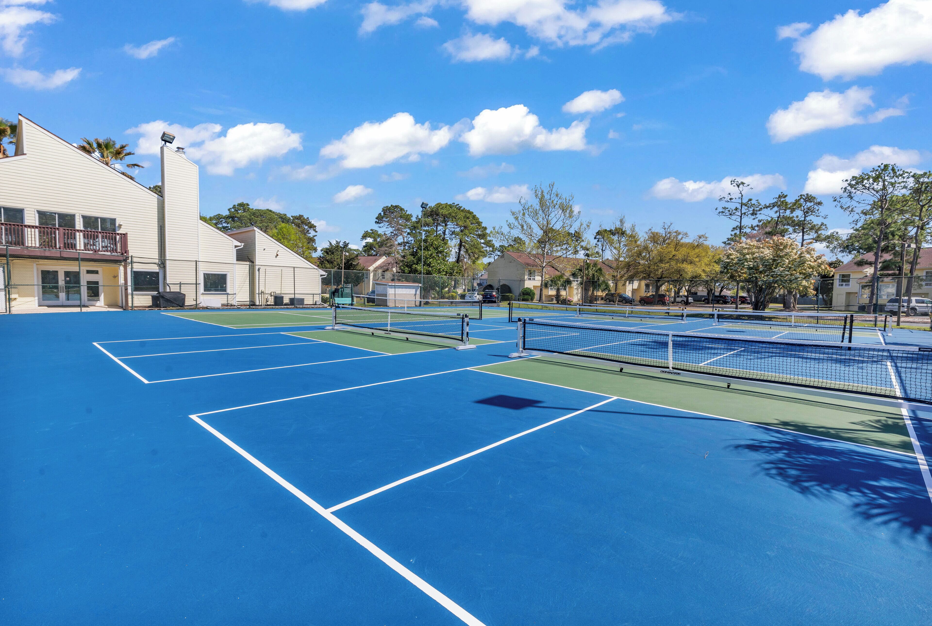A tennis court with multiple courts, a fence, and a building with a balcony in the background.