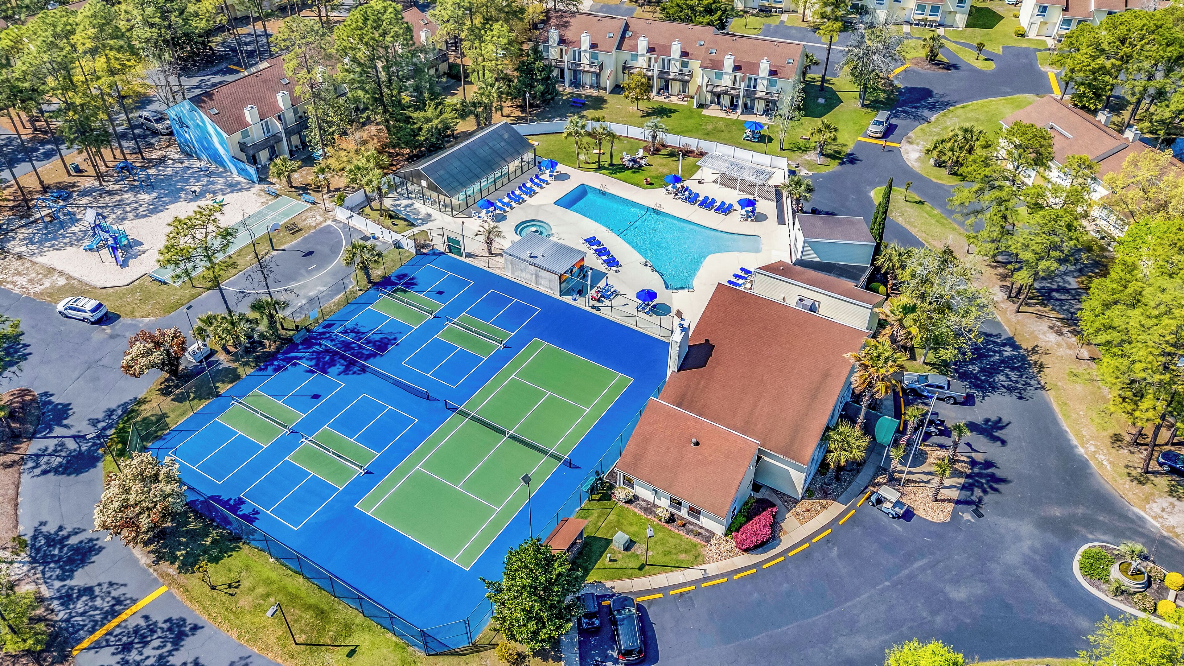 An aerial view of a residential area with a tennis court, swimming pool, and a small building with a red roof.