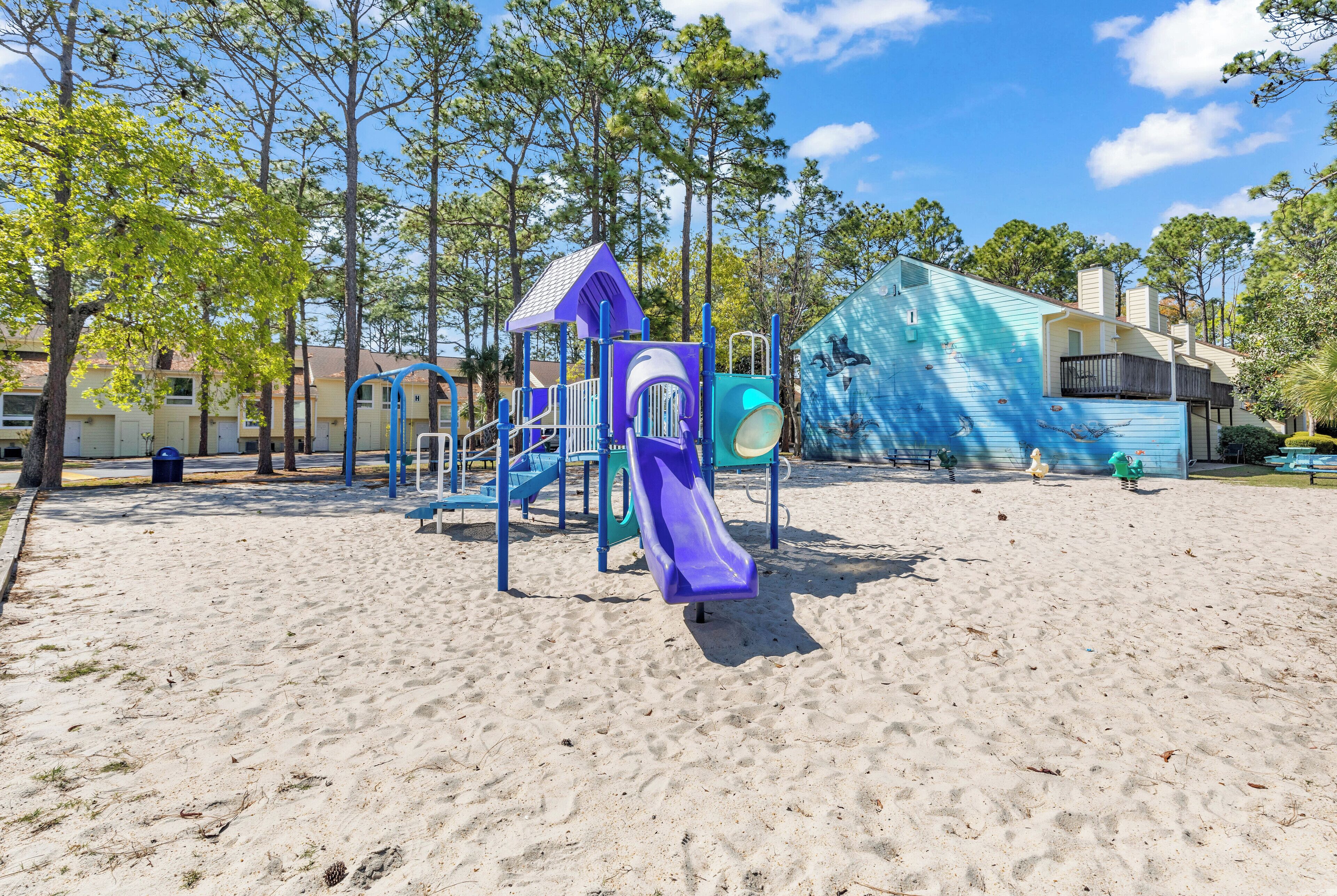 A playground with a purple slide and climbing structures, surrounded by trees and a building with a blue facade.