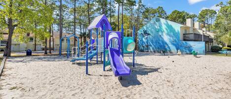 A playground with a purple slide and climbing structures, surrounded by trees and a building with a blue facade.
