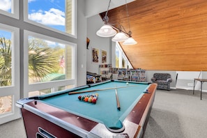 A pool table with balls, a pool cue, and a bookshelf with books and games.