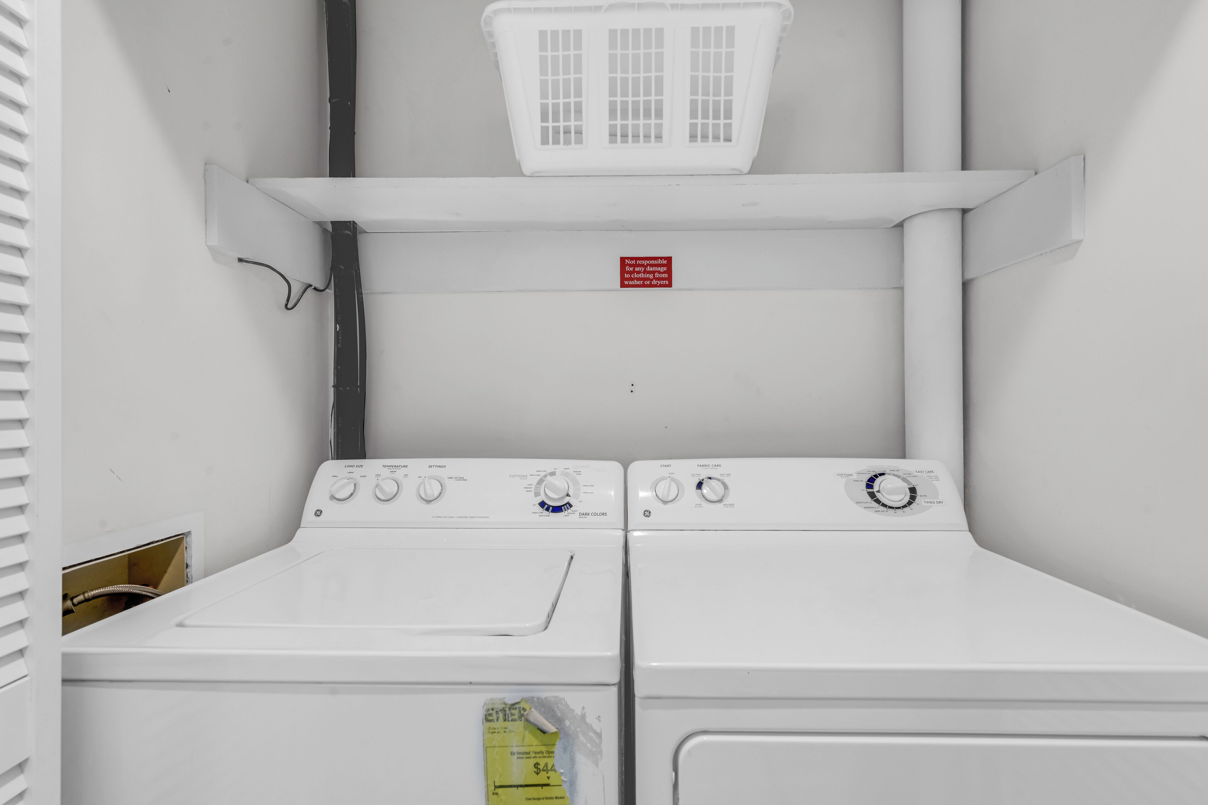 A laundry room with two stacked washers and dryers, a white basket on the shelf, and a warning sign.