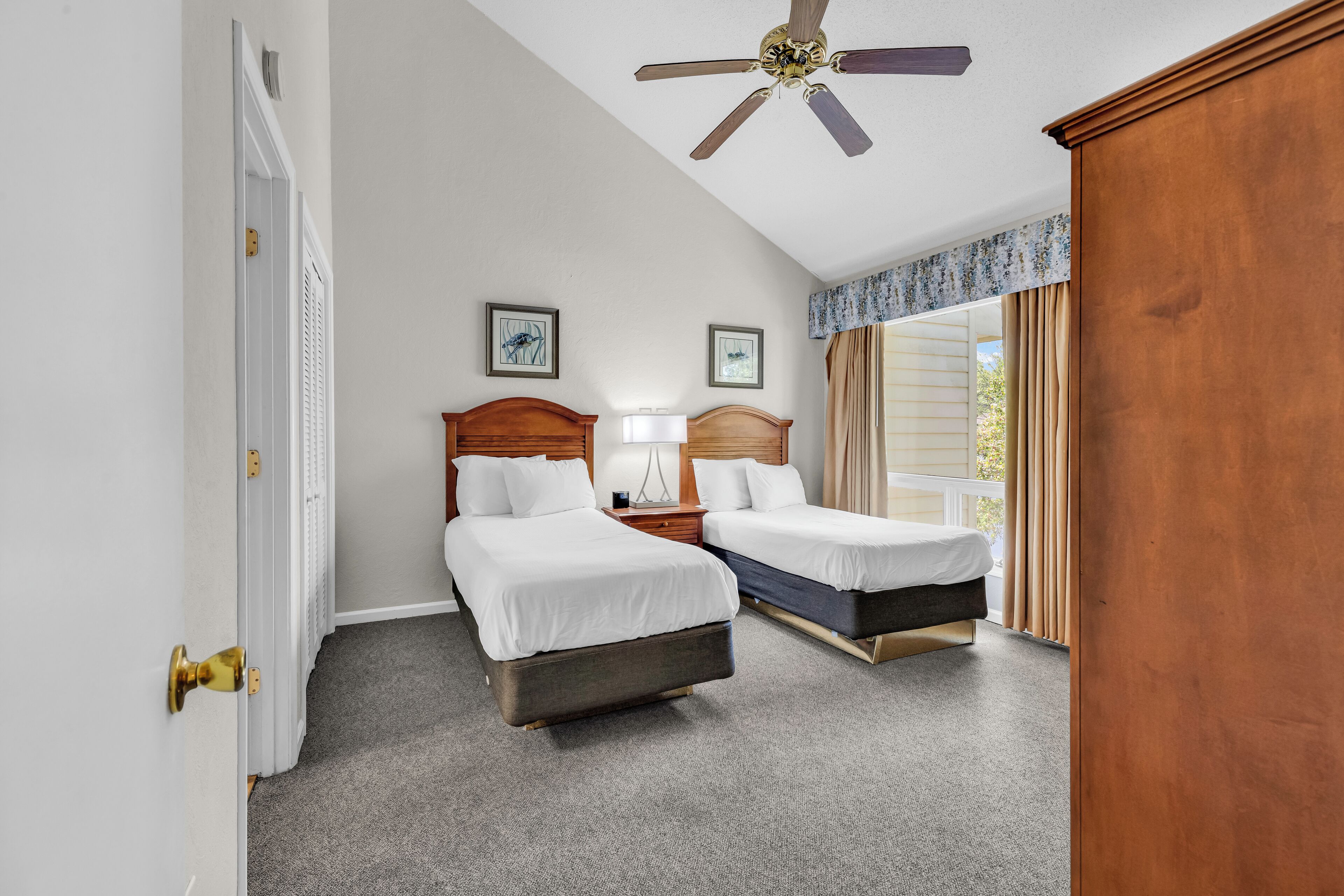 A hotel room with two beds, a ceiling fan, a window with curtains, and a door with a brass handle.