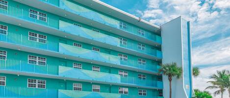 A multi-story hotel with a blue and white color scheme, featuring balconies and a parking area with several cars.
