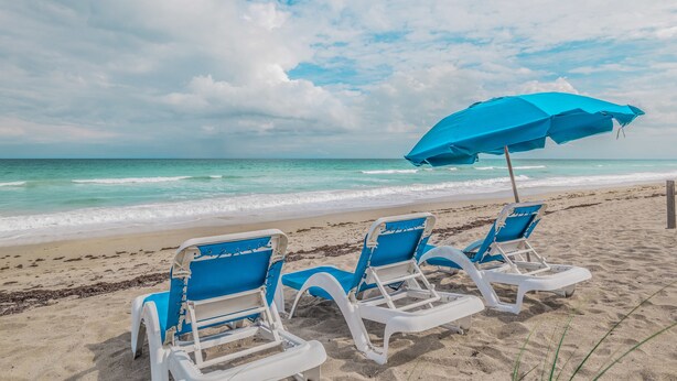Two beach chairs with blue cushions and a blue umbrella on a sandy beach with a view of the ocean. - Turtle Reef Club (Jensen Beach)