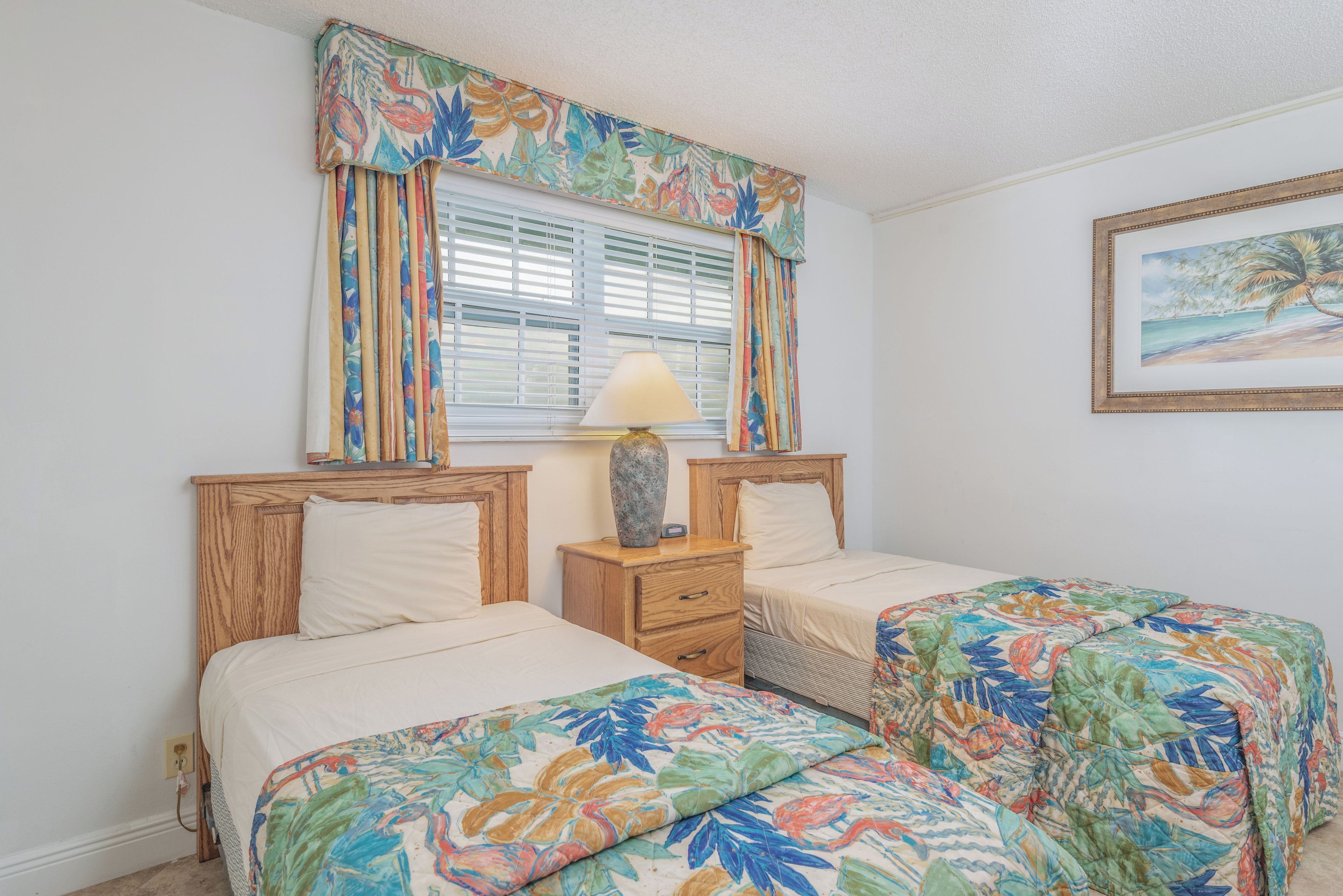 A bedroom with two single beds, a nightstand with a lamp, a window with blinds, and a painting on the wall.
