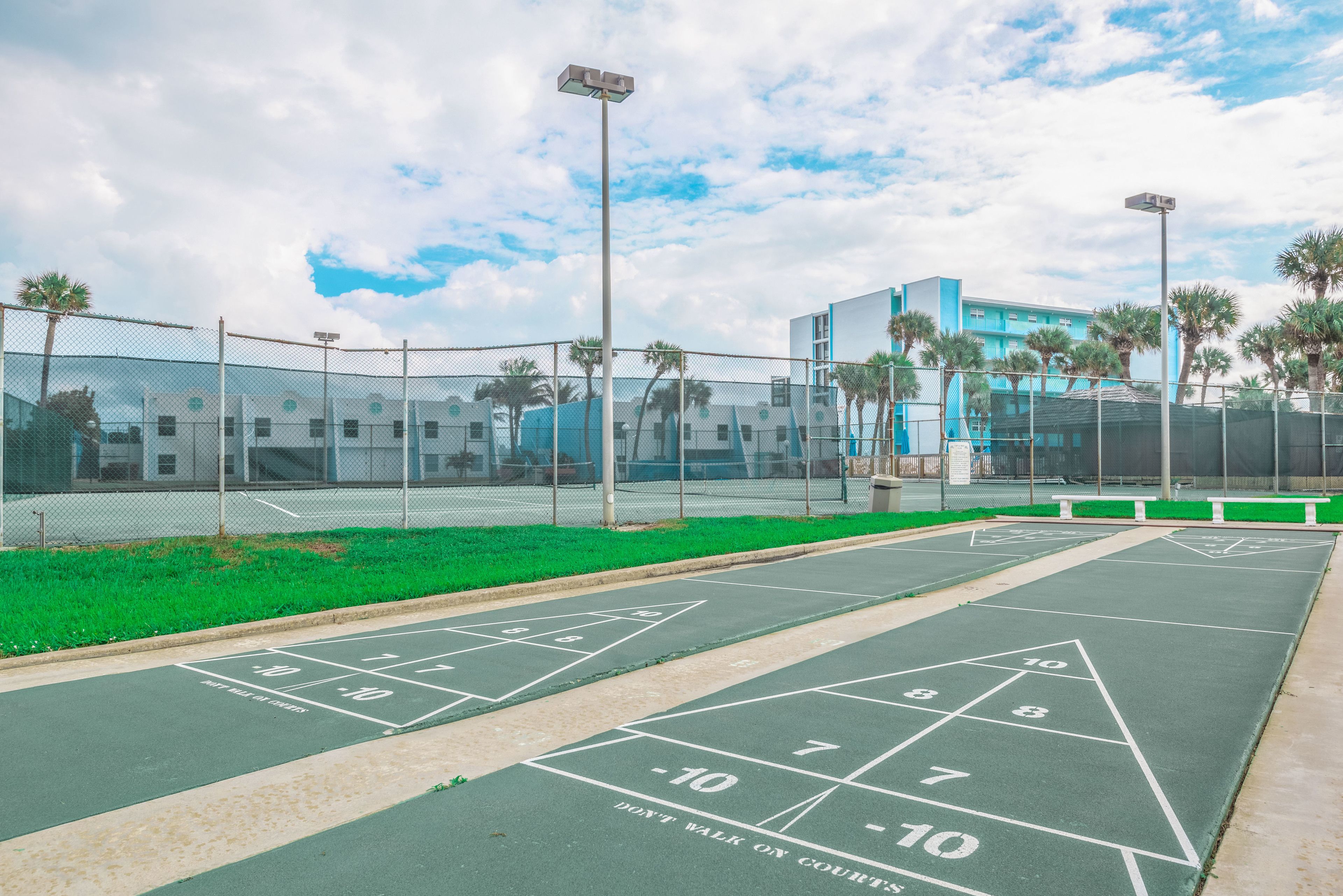 A shuffleboard court with numbers and "DON'T WALK ON COURTS" text, surrounded by a fence and palm trees, with a modern building in the background.