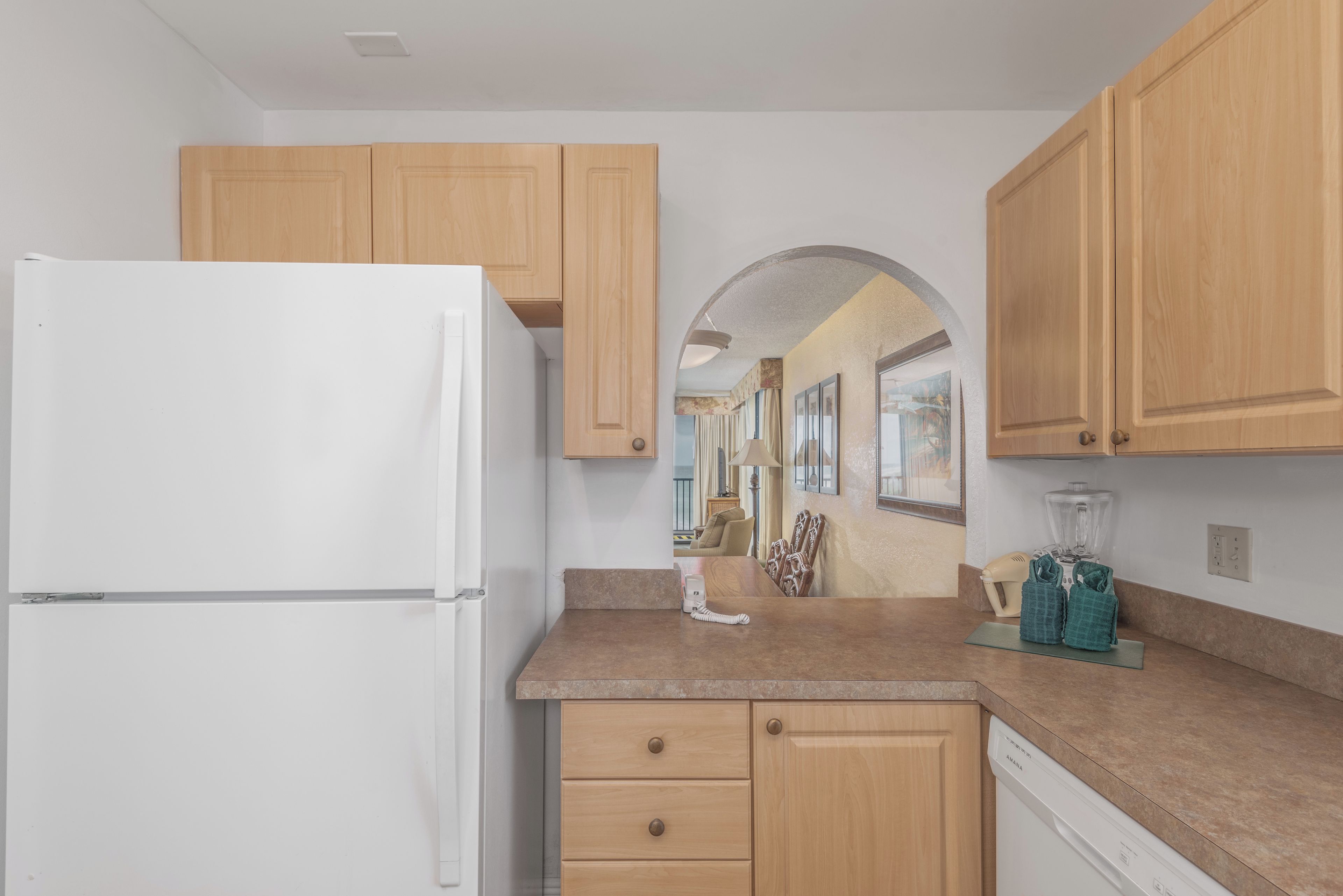 A kitchen with wooden cabinets, a white refrigerator, a countertop with a blender, and a view into the dining area.