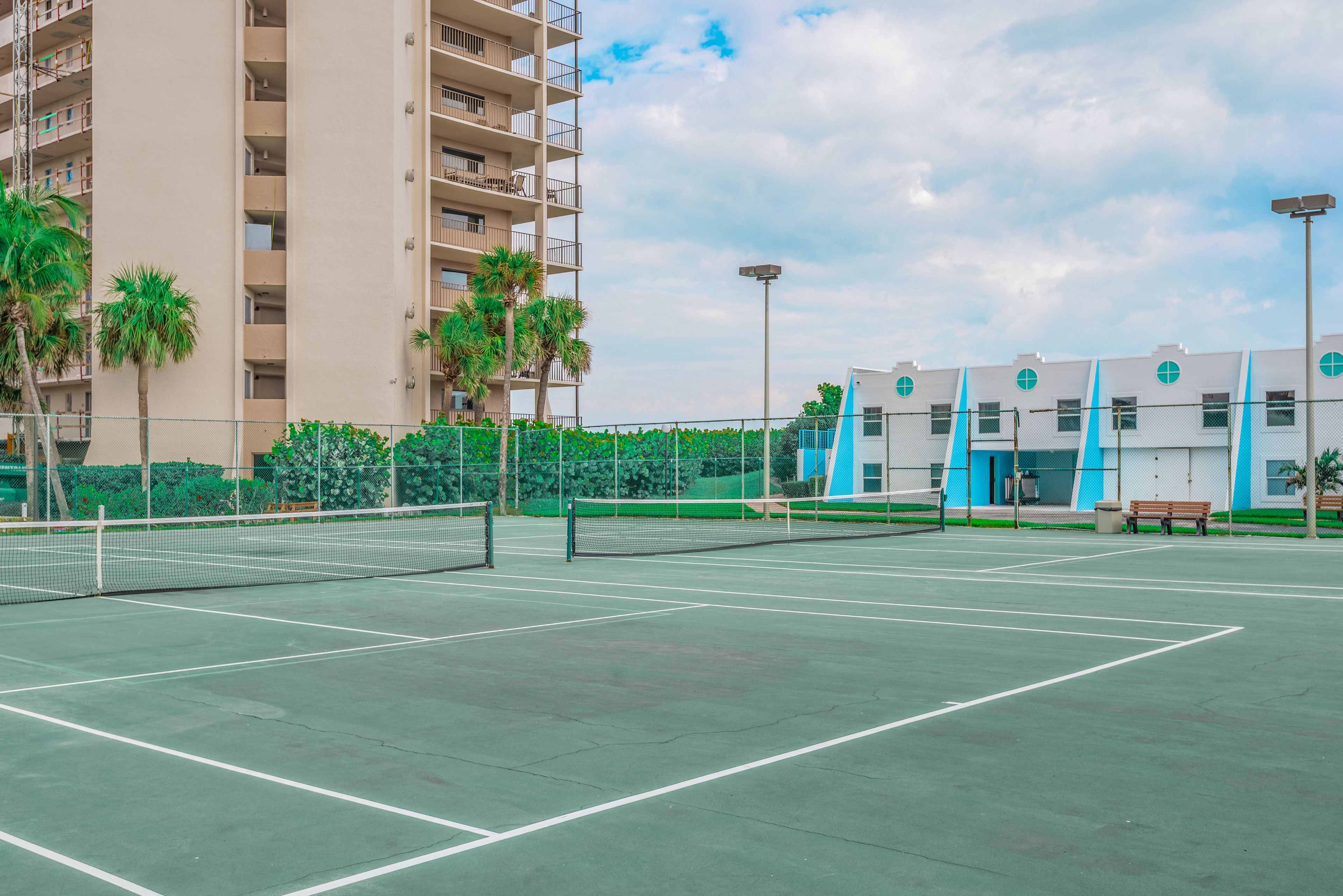 Un campo da tennis all'aperto con una rete, circondato da una recinzione, palme e edifici sullo sfondo.