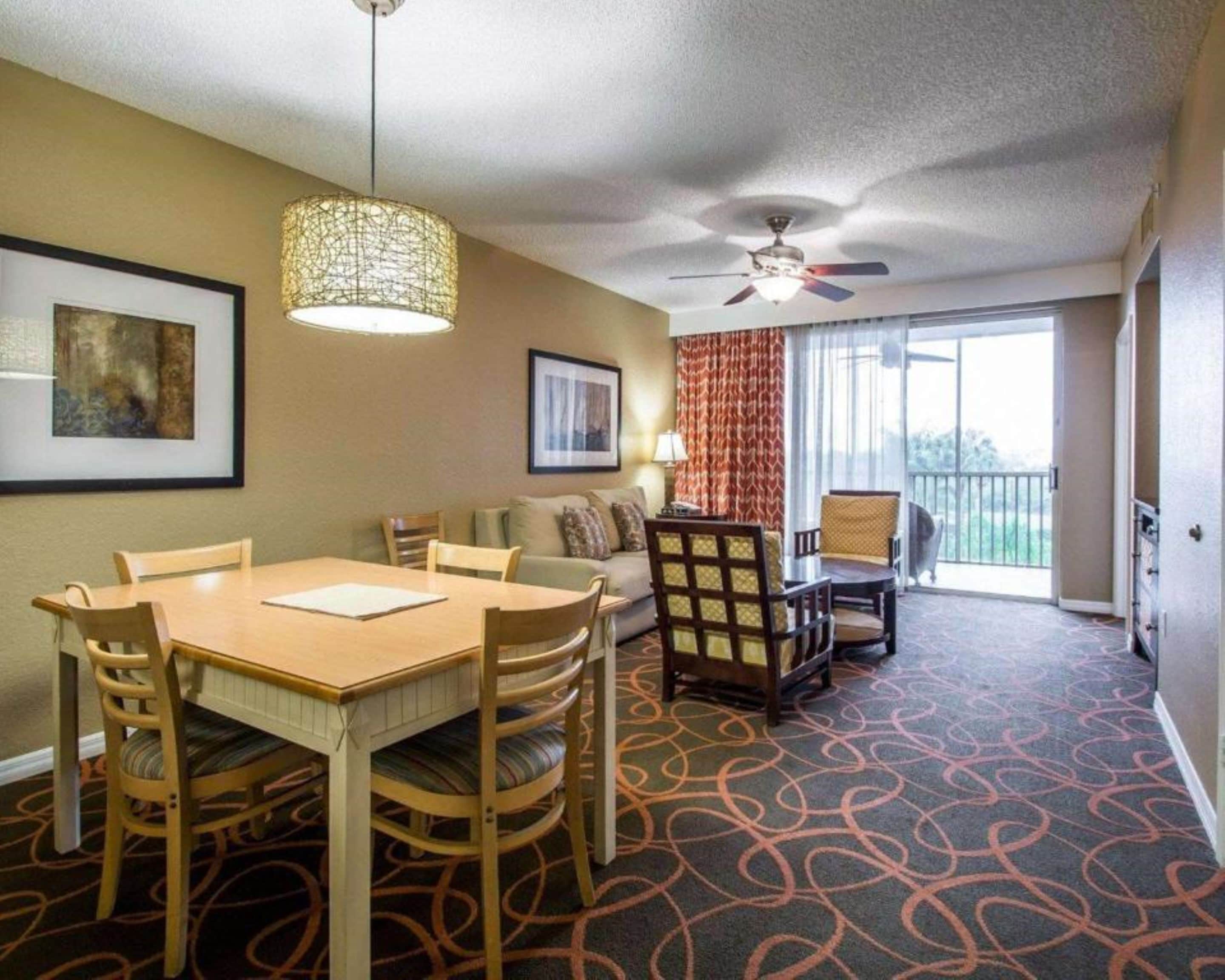 Une chambre d’hôtel équipée d’une table à manger, de chaises, d’un canapé, d’un ventilateur de plafond et d’une grande fenêtre avec des rideaux.