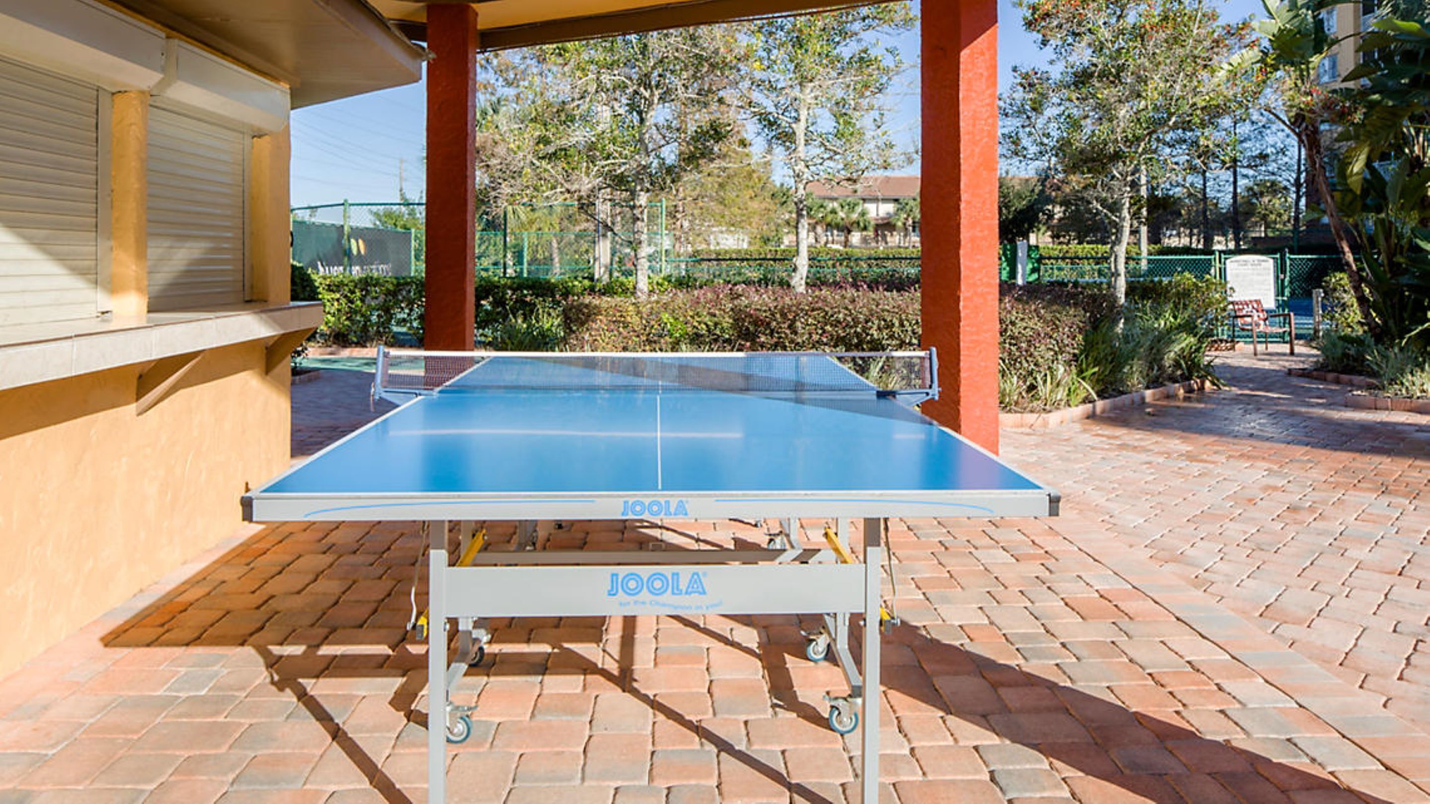 Et transportabelt ping pong-bord med hjul, mærket 'JOOLA', placeret på en fliseterasse med et overdækket område og grøn bevoksning i baggrunden.