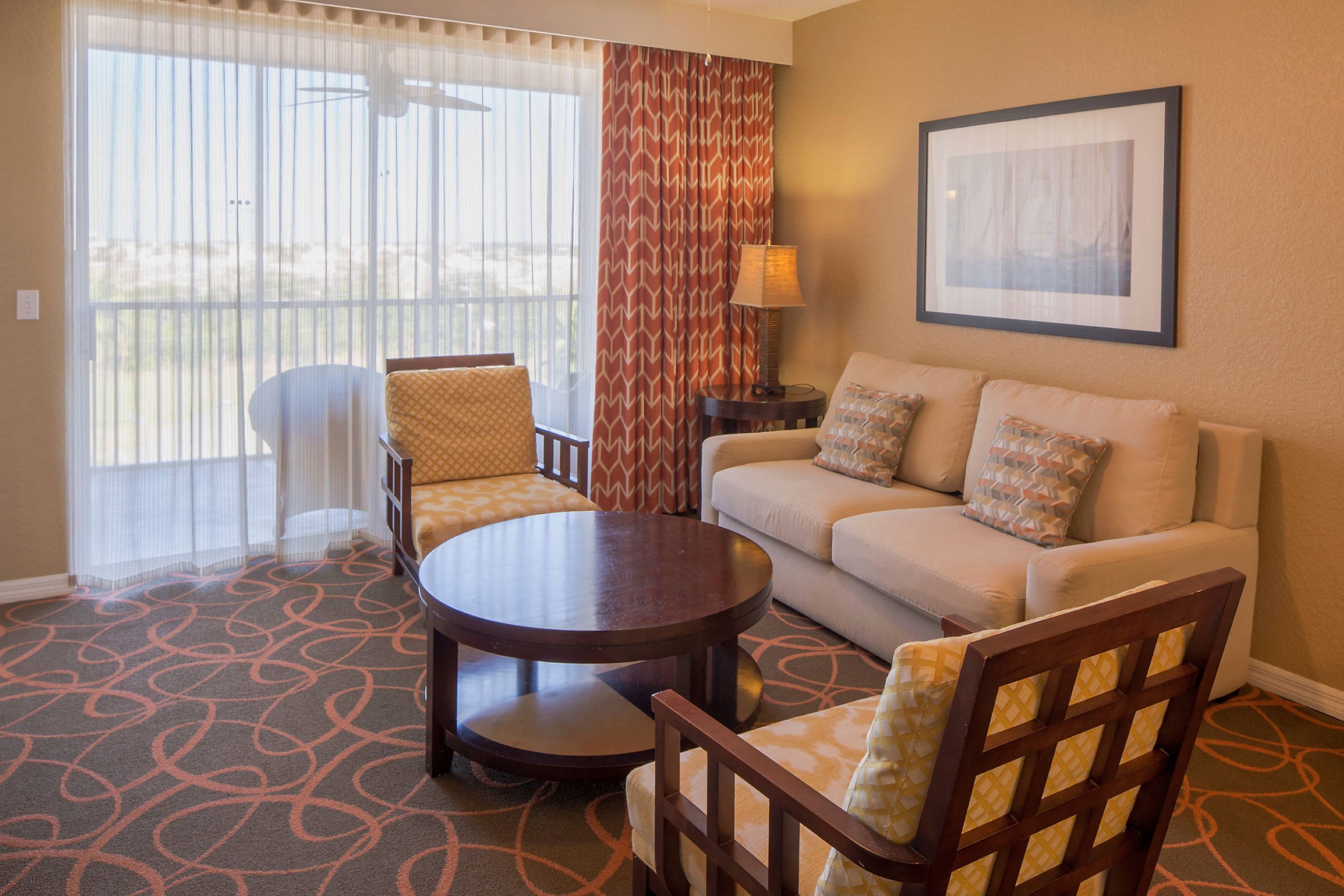 A hotel room with a sofa, two armchairs, a coffee table, a lamp, a framed picture on the wall, and a view through the window.