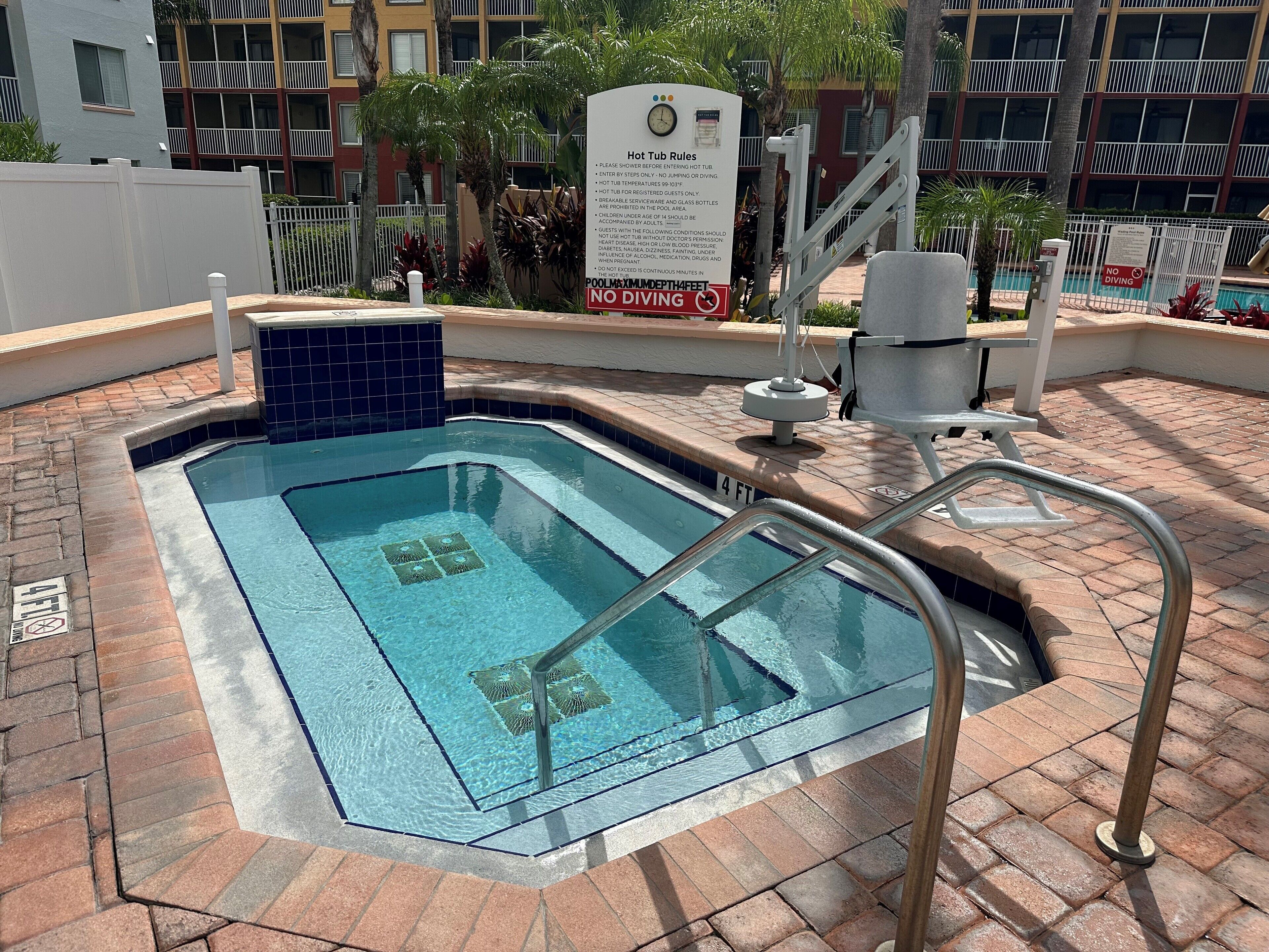 Un bain à remous agrémenté d’un panneau indiquant les règles d’utilisation, une échelle pour la piscine et un espace au bord de la piscine doté d’un sol en briques et d’un panneau « Interdiction de plonger ».