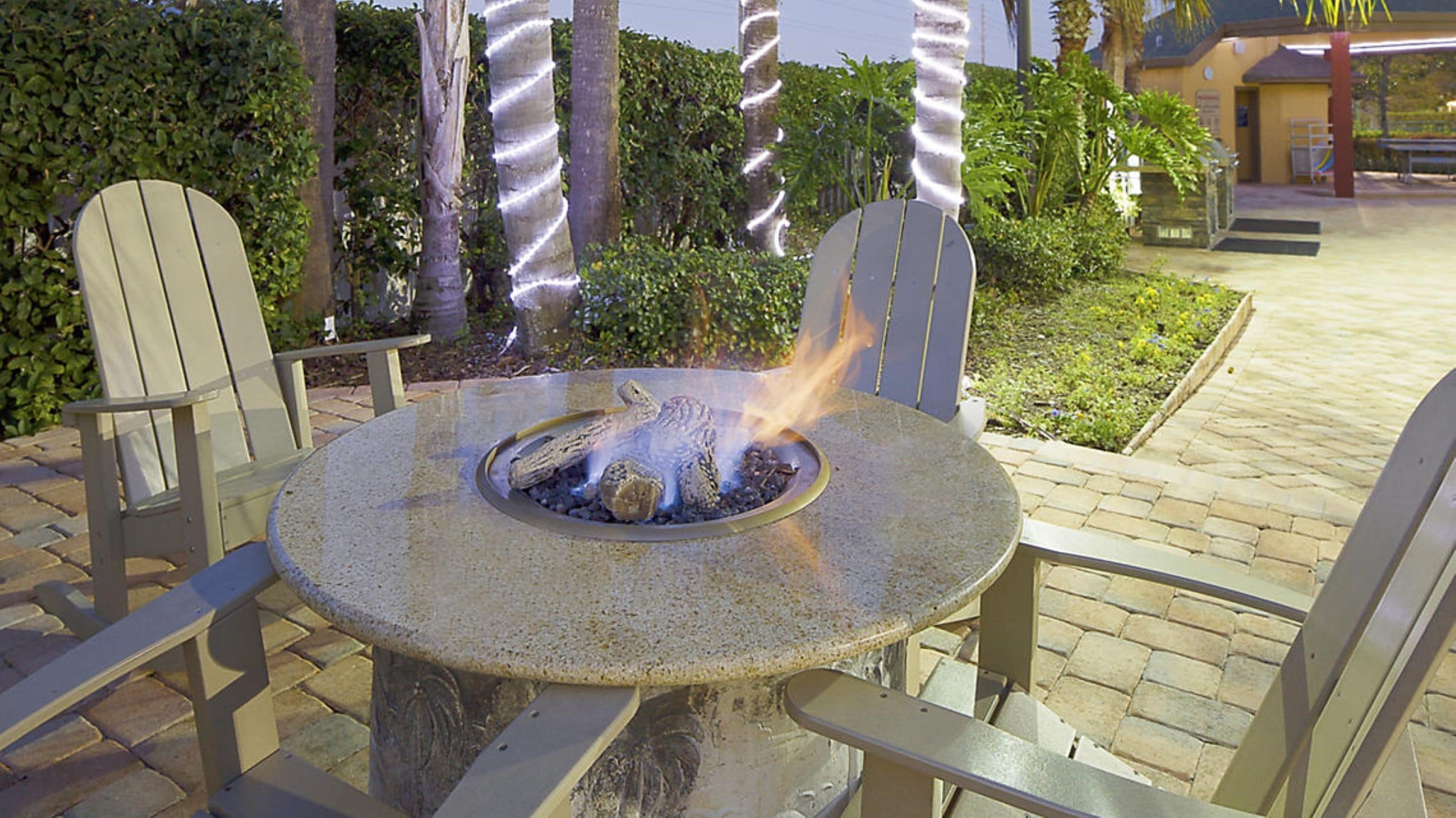 A round stone table with a fire pit in the center, surrounded by two Adirondack chairs.