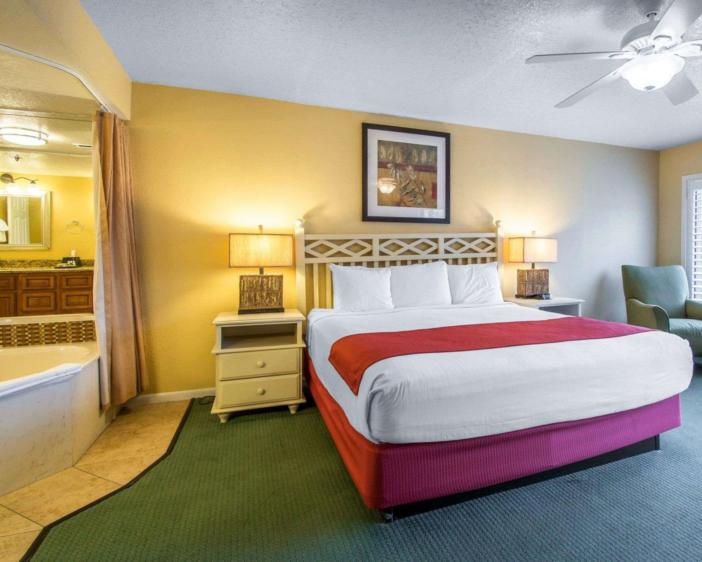 A hotel room with a bed, a nightstand, a lamp, a ceiling fan, a bathroom with a bathtub, and a painting on the wall.