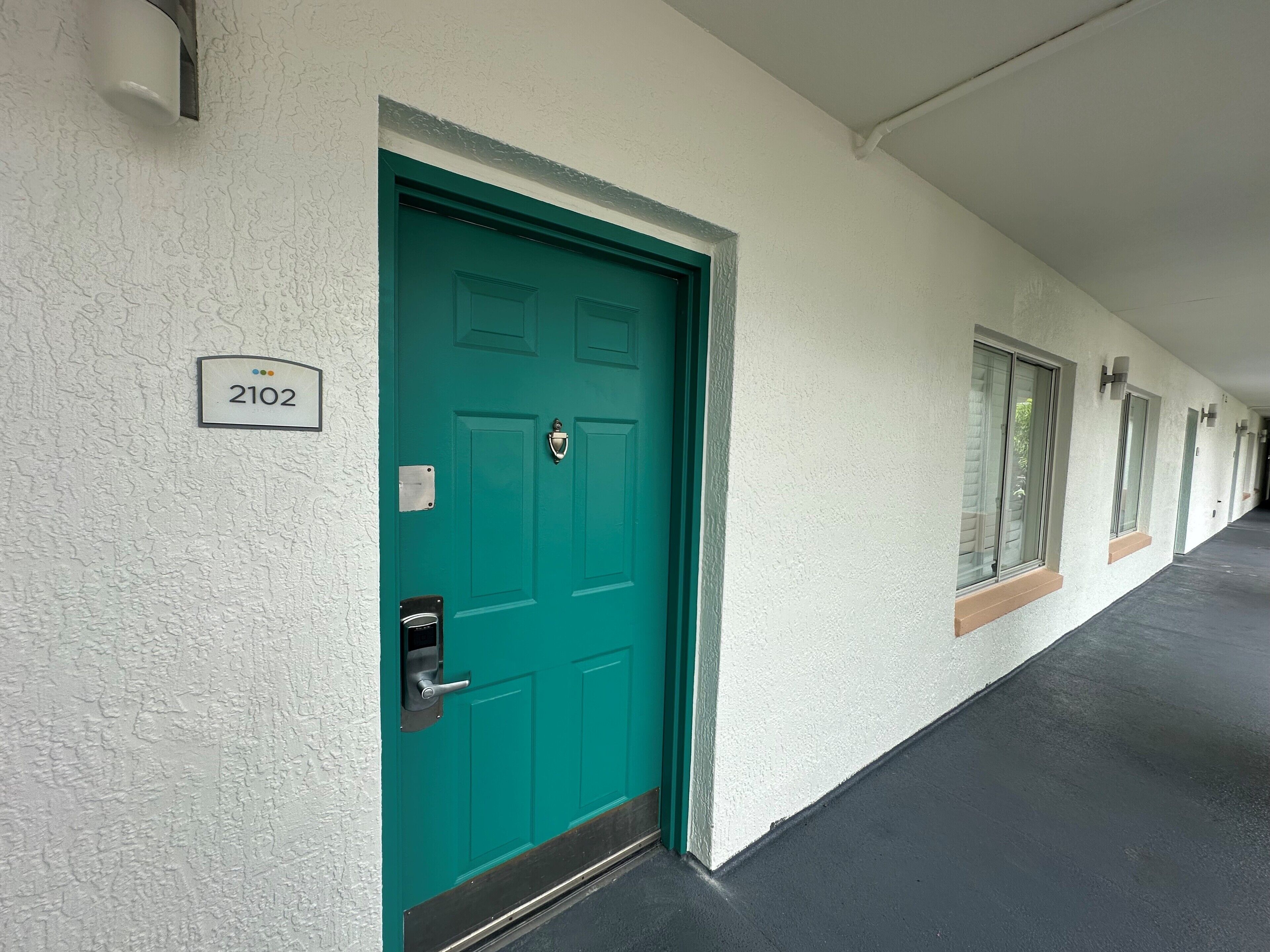 Une porte de couleur turquoise, ornée d’une plaque portant le numéro « 2102 » et d’un trou de serrure, faisant partie d’un immeuble à la façade blanche et au plancher foncé.