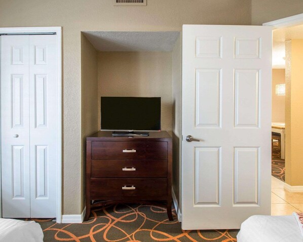 A hotel room with a closed door, a dresser with drawers, a television on top, and a carpet with a geometric pattern.