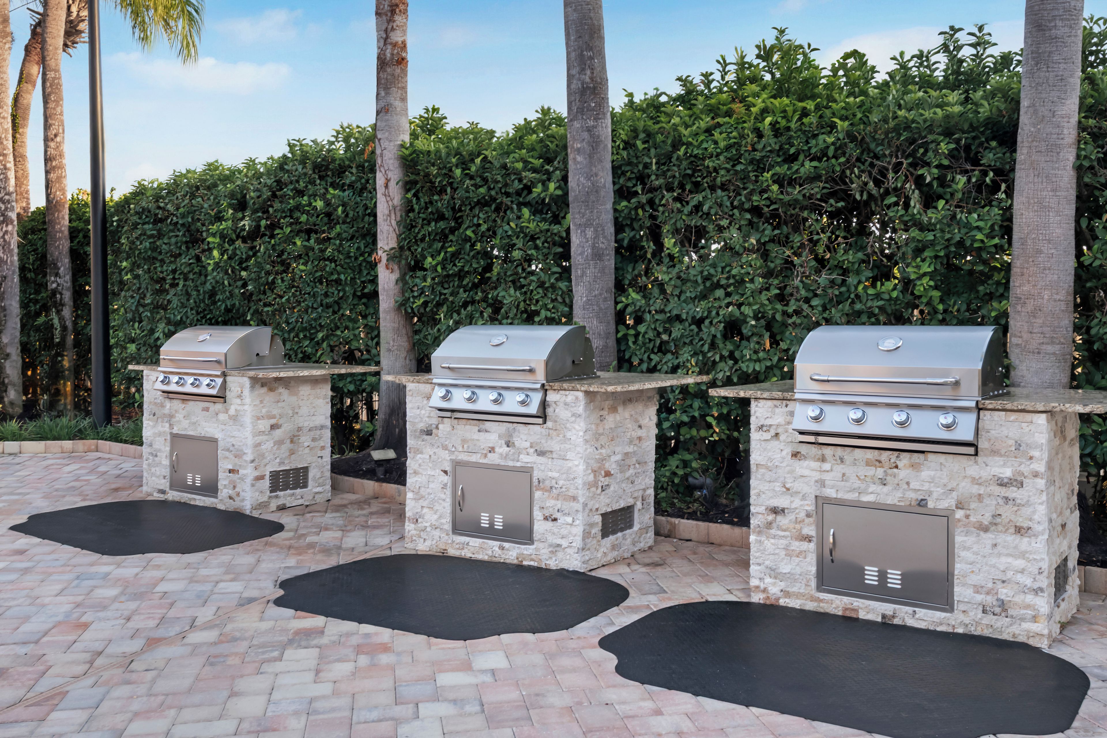 Three outdoor barbecue grills with stone bases, set on a paved patio with black mats.