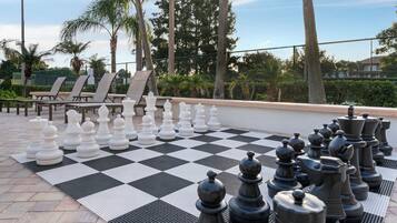 Un jeu d’échecs installé sur une terrasse, avec des fauteuils et des palmiers en arrière-plan.