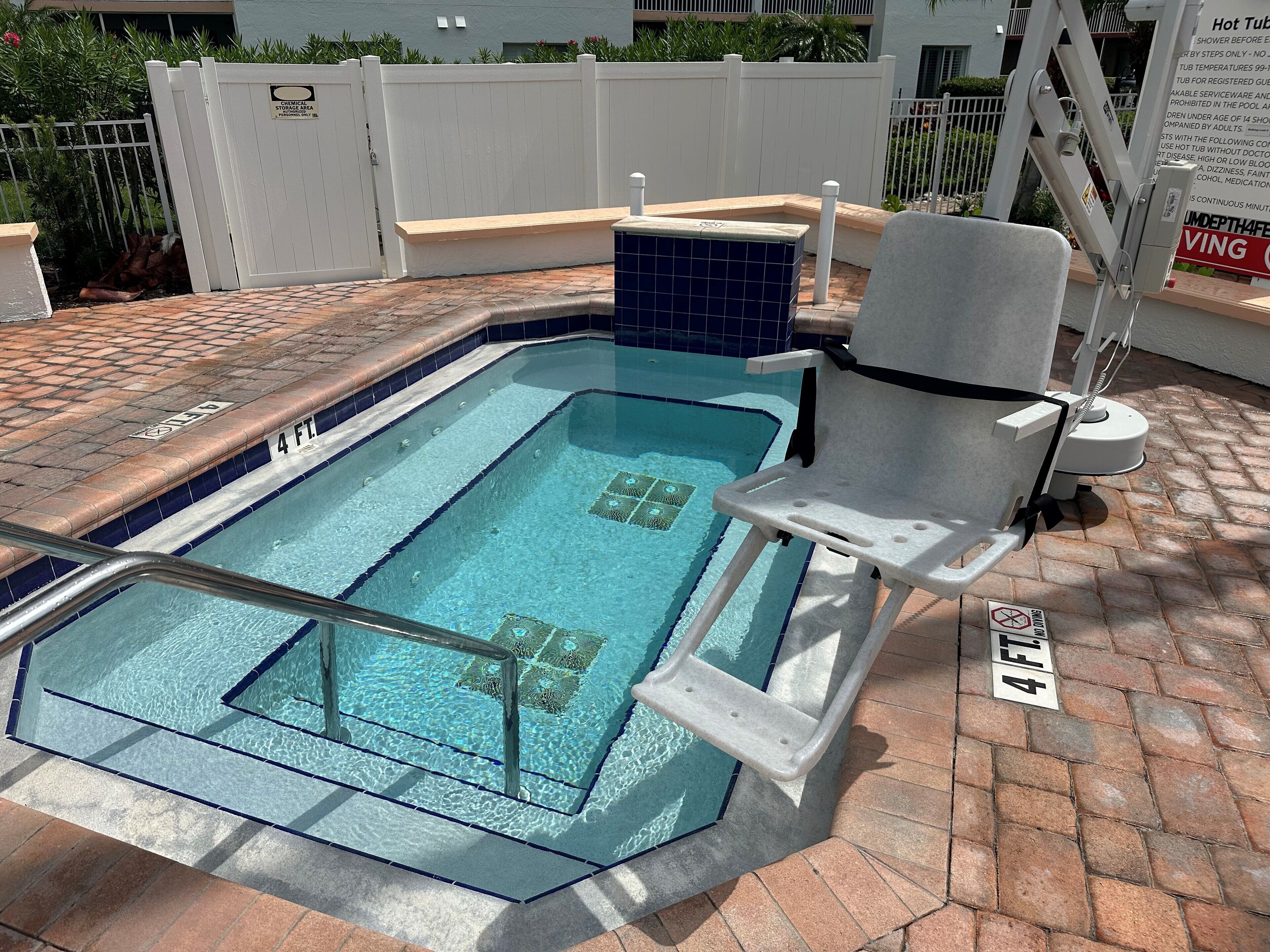 Una vasca idromassaggio con una panca e una sedia a bordo piscina, circondata da un patio in mattoni e un recinto bianco.