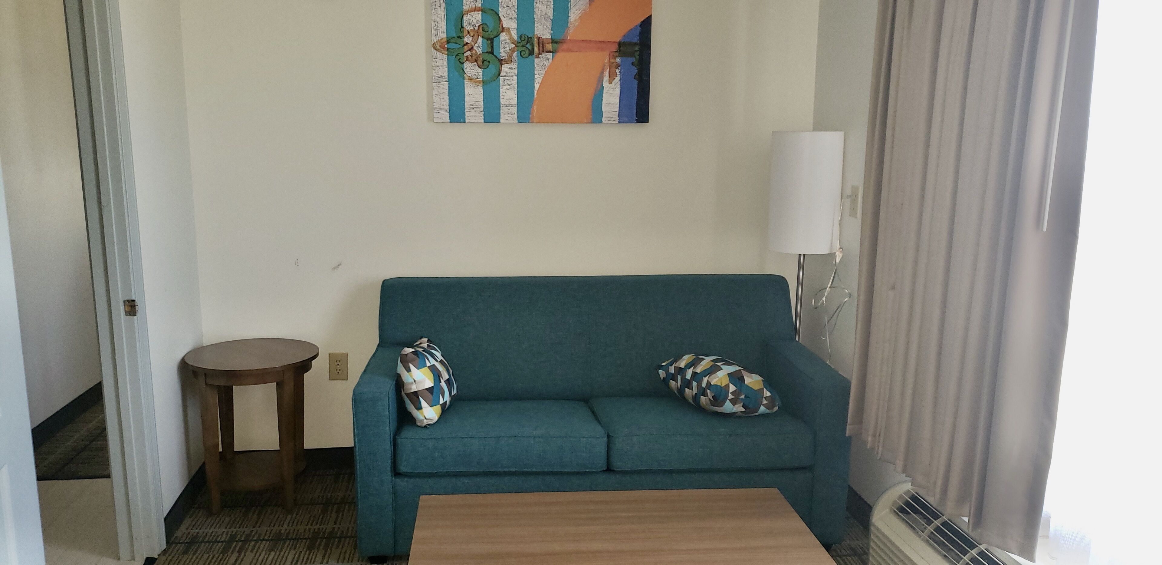 A teal sofa with patterned pillows, a wooden side table, a painting on the wall, and a white lamp.
