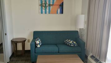 A teal sofa with patterned pillows, a wooden side table, a painting on the wall, and a white lamp.