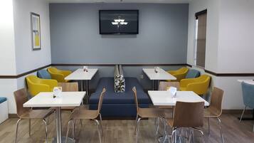 A modern hotel room with a blue sofa, yellow and blue chairs, white tables, and a flat-screen TV on the wall.