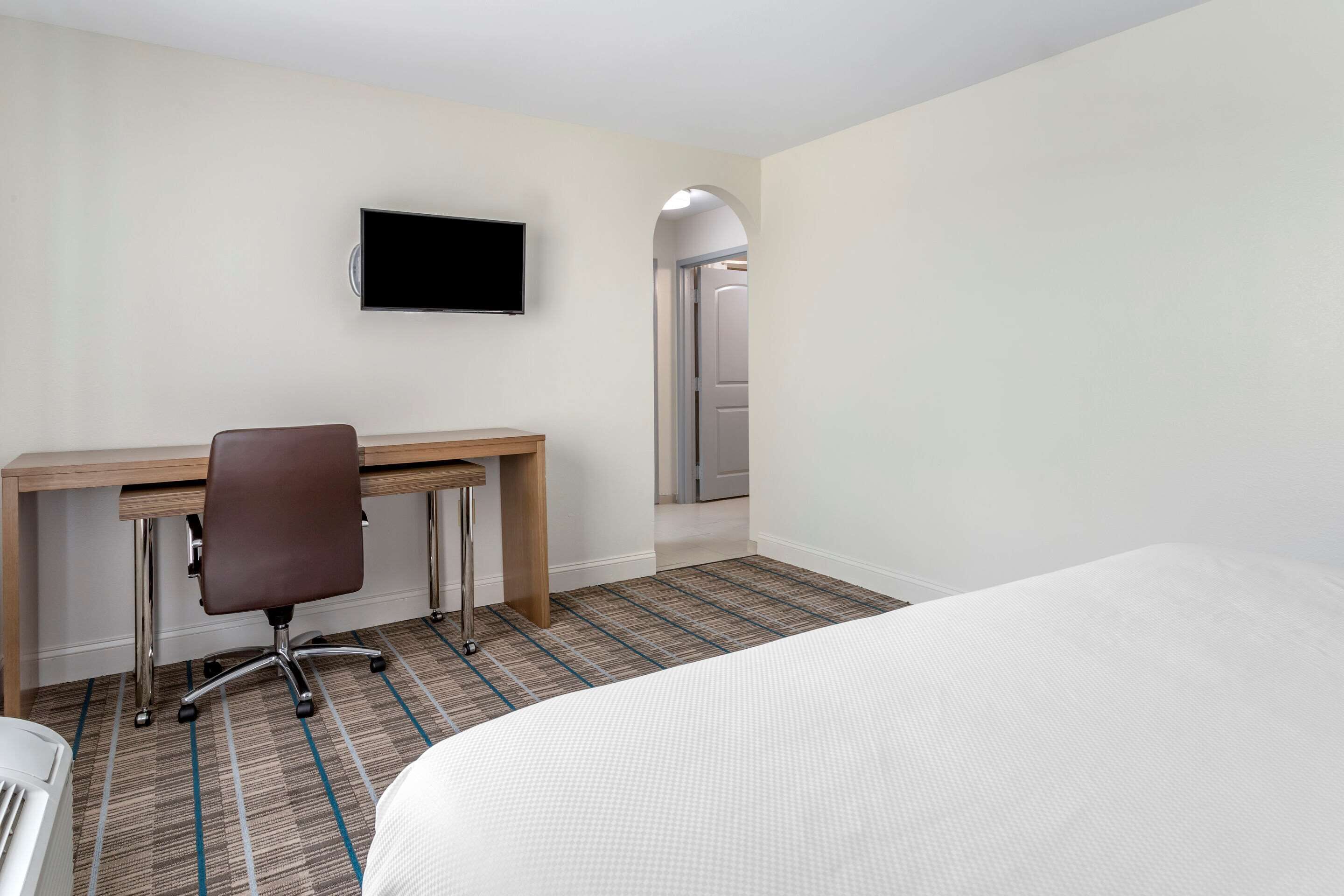 Habitación de hotel con cama, escritorio con silla, televisor de pantalla plana y puerta que da a un pasillo.