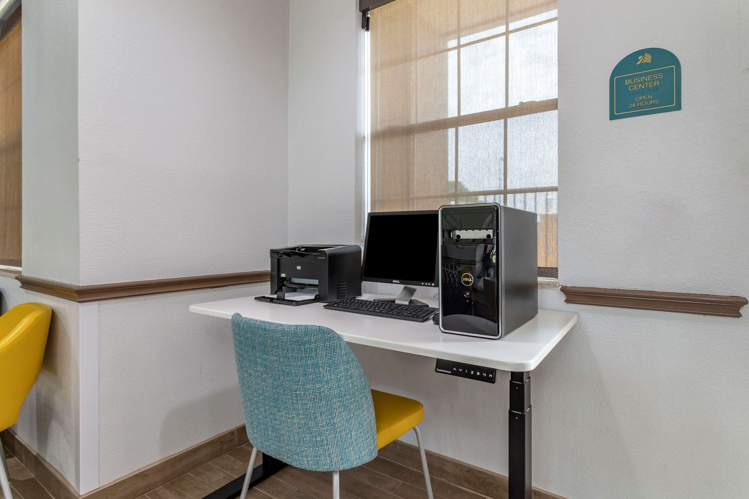 A corner office with a desk, two monitors, a printer, and a chair. A sign indicates a business center open 24 hours.