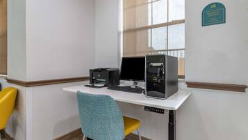 A corner office with a desk, two monitors, a printer, and a chair. A sign indicates a business center open 24 hours.