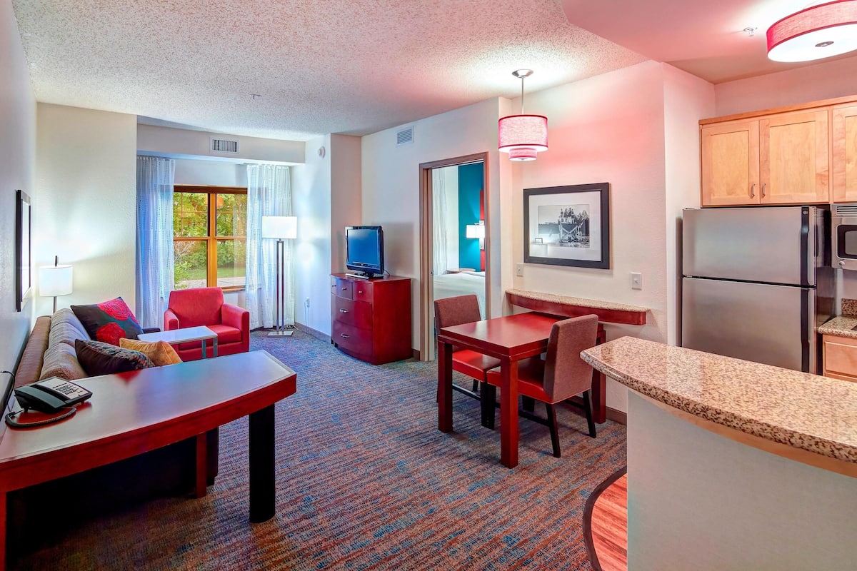 A hotel room with a living area, a dining table, a TV, a refrigerator, and a kitchen area.