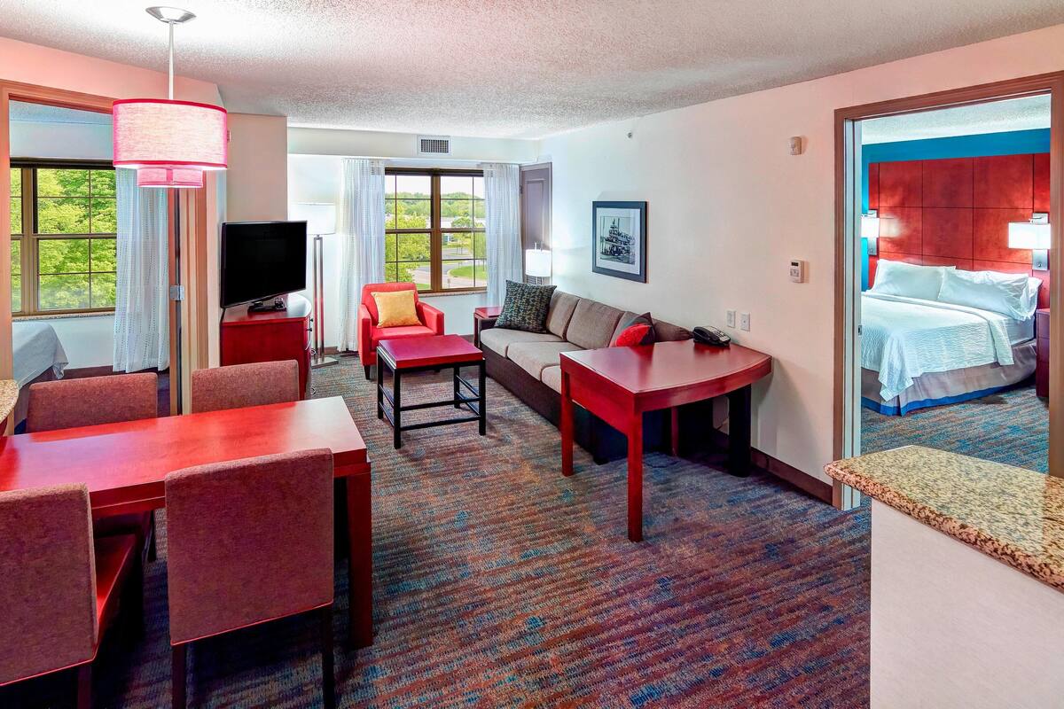 A hotel room with a dining area, a sofa, a TV, and a bed with a view of the outdoors.
