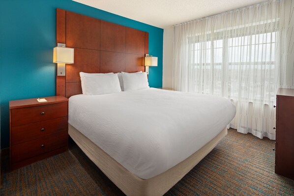 A hotel room with a large bed, wooden headboard, two bedside tables with lamps, a nightstand, and a window with sheer curtains.