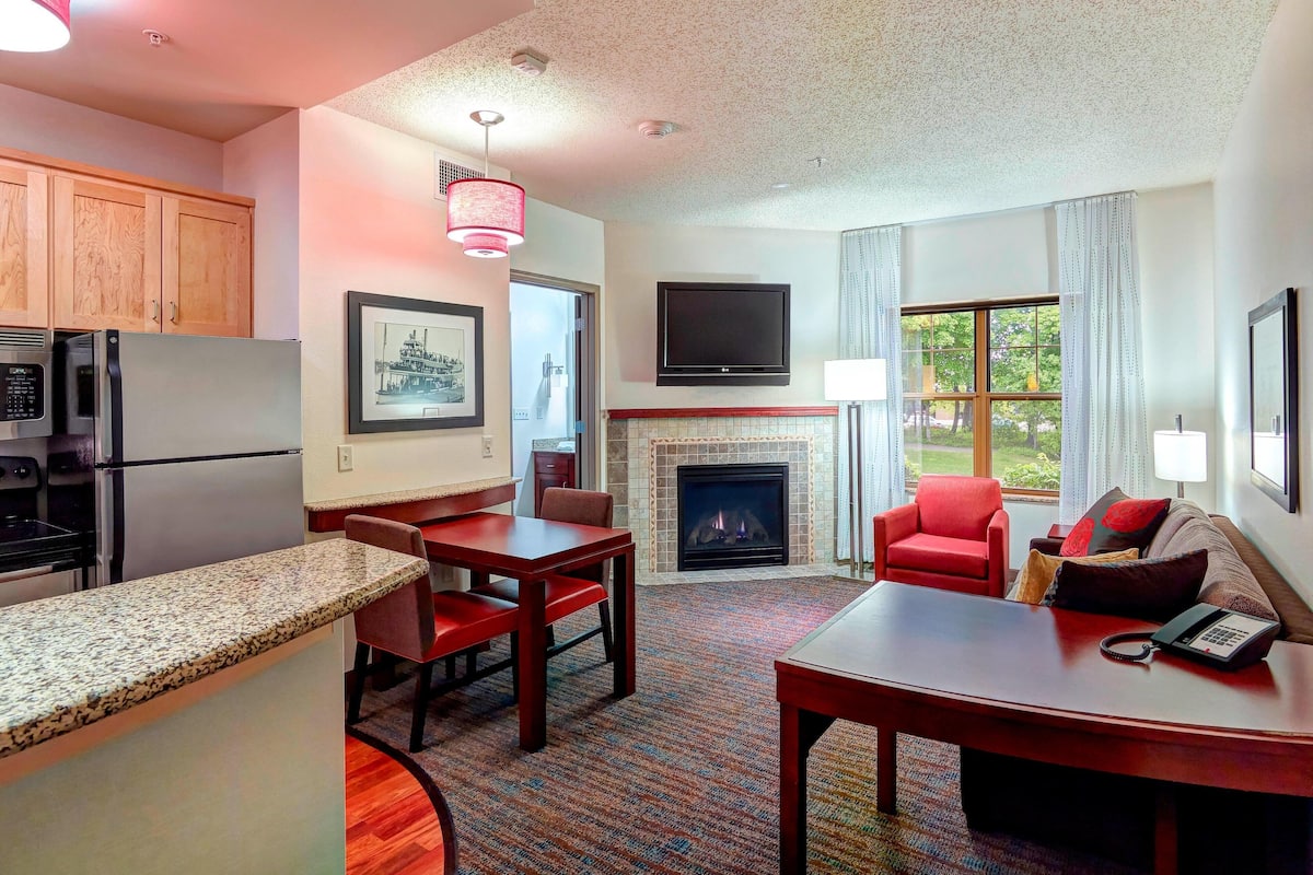 A modern kitchen with granite countertops, a dining area with a red chair, a fireplace, and a TV mounted on the wall.