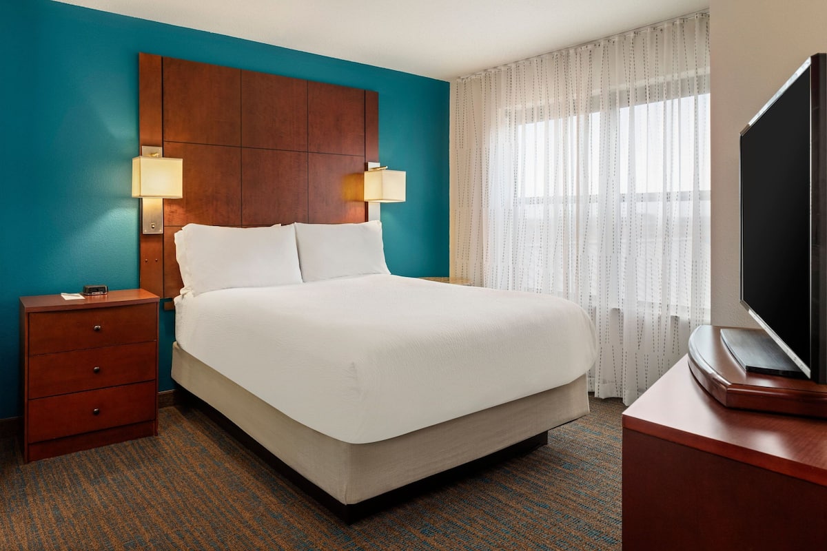 A hotel room with a large bed, a wooden headboard, two bedside lamps, a flat-screen TV, and a window with sheer curtains.