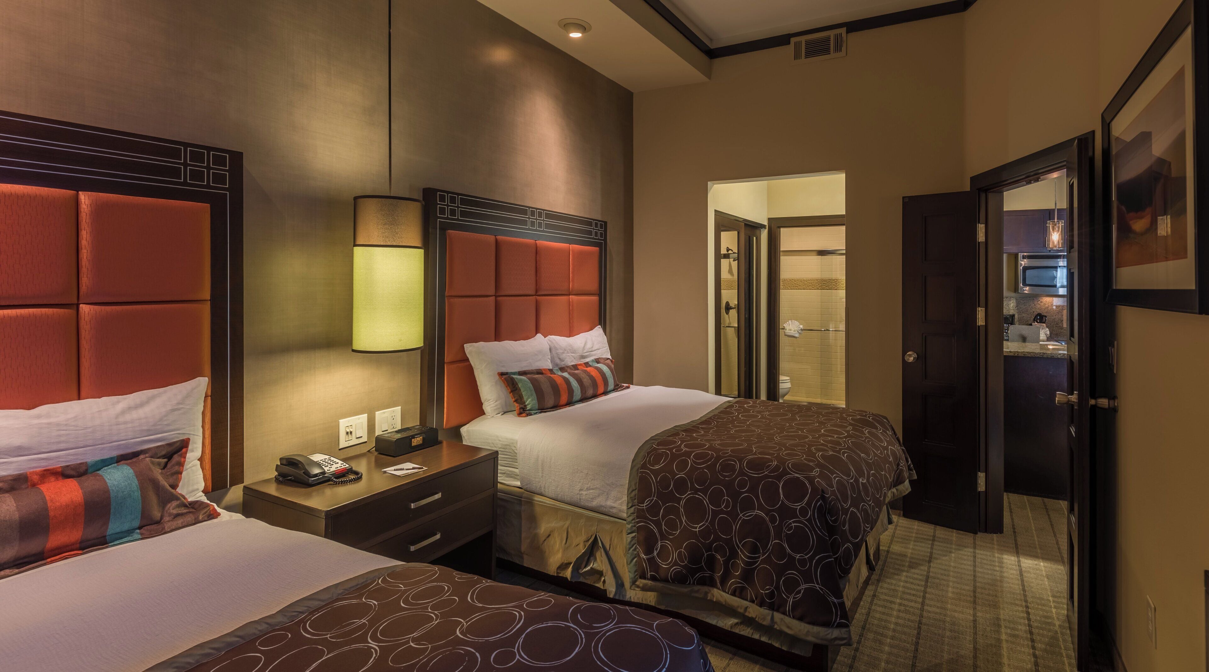 A hotel room with two beds, a bedside table with a lamp, a bathroom visible through an open door, and a kitchenette area.