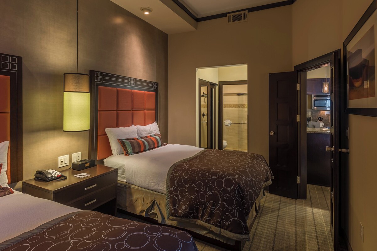 A hotel room with two beds, a bedside table with a phone, a lamp, and a bathroom visible through an open door.