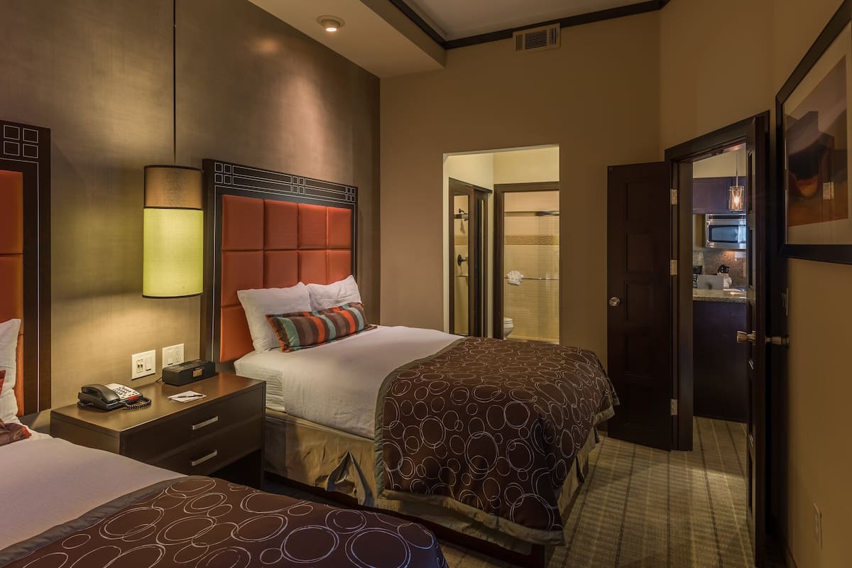 A hotel room with two beds, a bedside table with a phone, a lamp, and a bathroom visible through an open door.