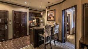 A modern kitchen with dark wood cabinets, a stainless steel refrigerator, a microwave, and a built-in oven. There are two bar stools with patterned cushions, a small island with a sink, and a hanging light fixture. - Staybridge Suites DFW Airport North by IHG (Irving)