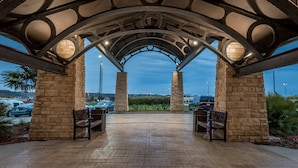 An outdoor covered patio with stone pillars, benches, and a view of parked cars and greenery. - Staybridge Suites DFW Airport North by IHG (Irving)