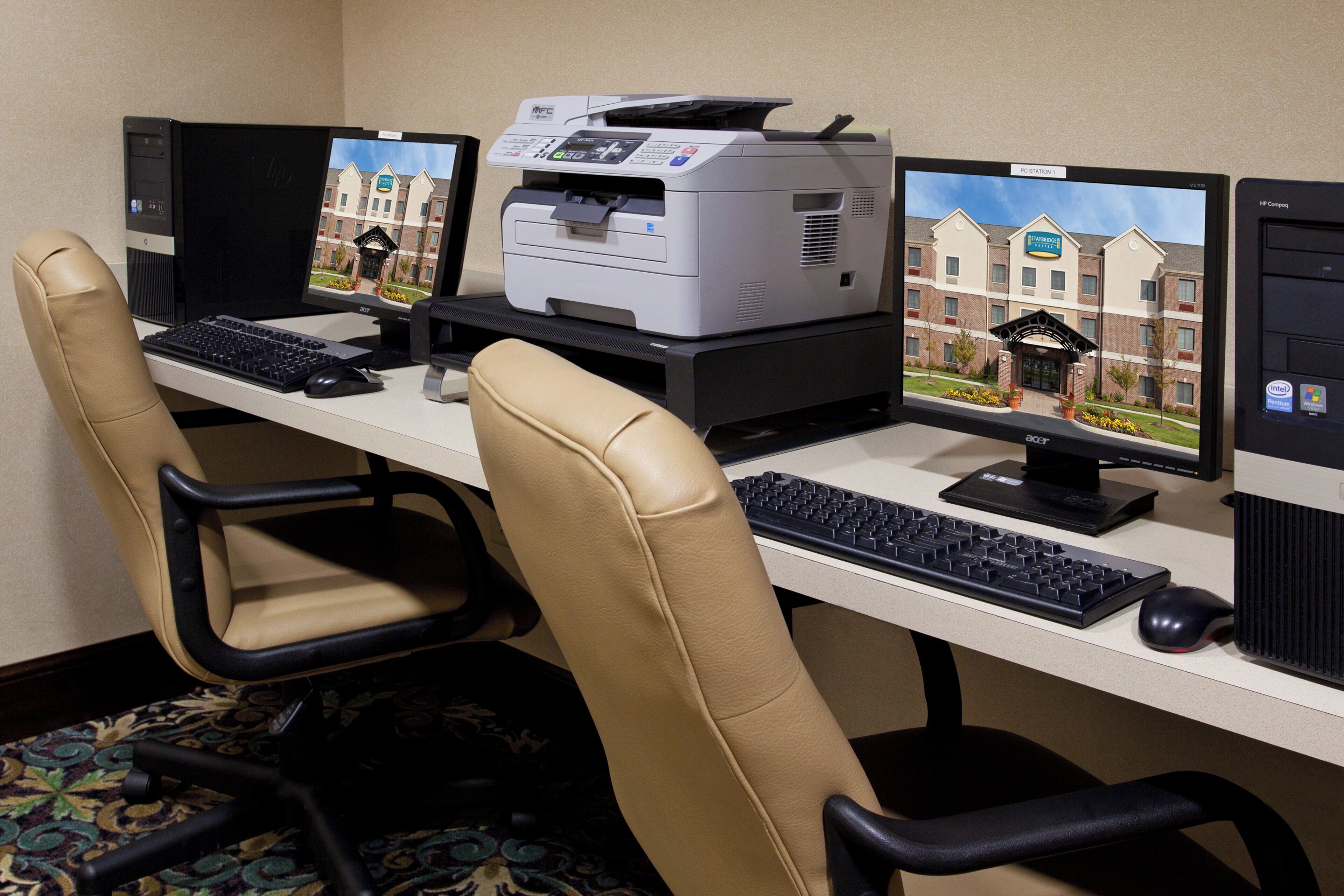 A hotel room with a desk equipped with two monitors, a printer, and a keyboard.